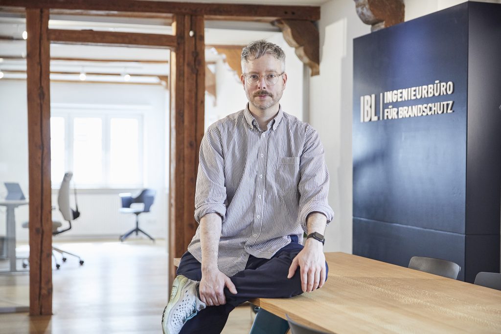Benedikt von IBL Ingenieurbüro für Brandschutz GmbH sitzt auf einem Schreibtisch in den Büroräumen in Hamburg