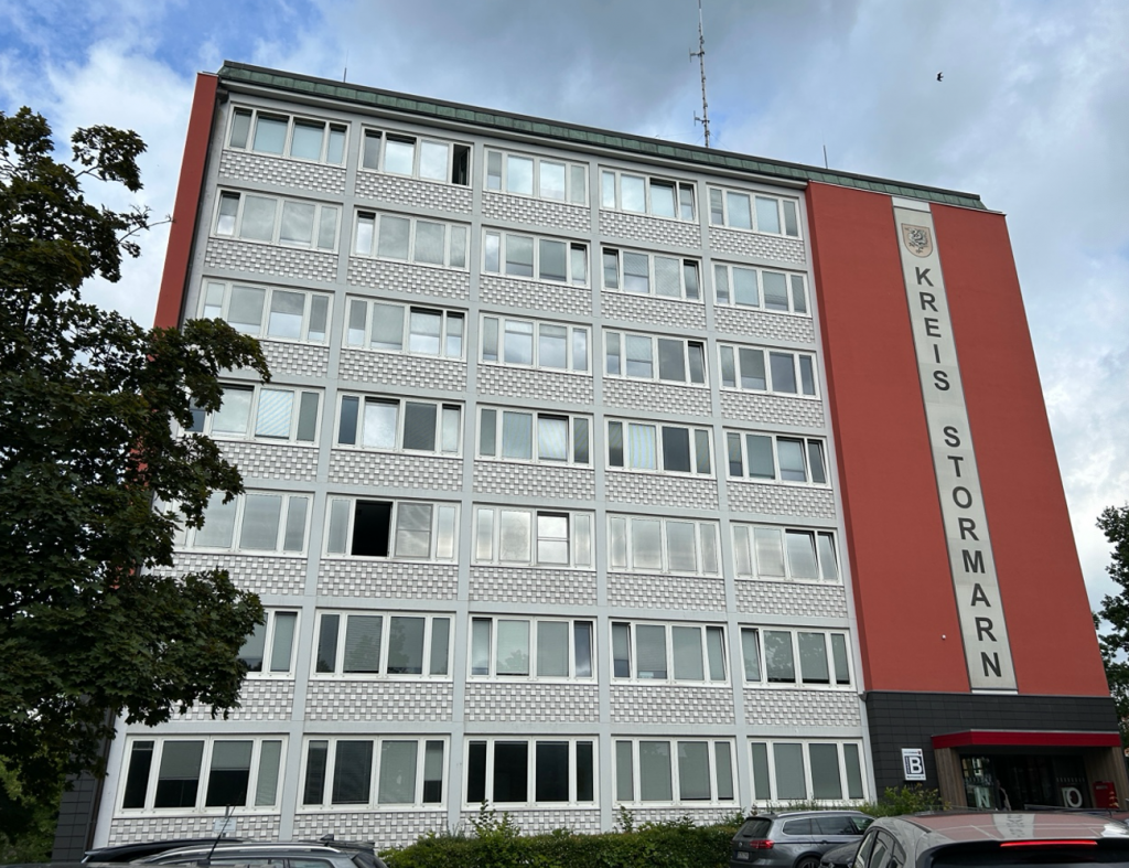 Sechsstöckiges Bürogebäude mit rot-weißer Fassade und dem Schriftzug Kreis Stormarn; Ingenieurbüro für Brandschutz, davor Parkplätze.